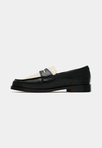 Mocassim de couro preto e bege com um salto baixo e um detalhe de tira na parte superior, mostrado de lado sobre um fundo branco.