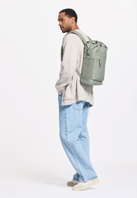 Vihreä kangasreppu, jossa on yläkahvat ja säädettävät olkahihnat. Repussa on etupuolella vetoketjullinen tasku ja värikkäät vetoketjunvetimet.