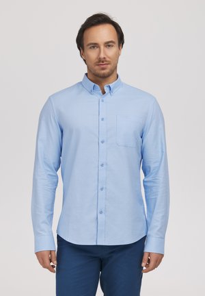 Mężczyzna stojący przodem, ubrany w jasnoniebieską koszulę na guziki z kieszenią na piersi oraz ciemnoniebieskie spodnie.