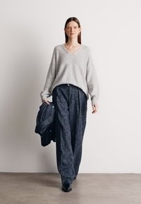 Pull de lã cinza claro com decote em V, combinado com calças largas de padrão escuro e uma jaqueta azul-marinho pendurada em um braço. Sapatos pretos.