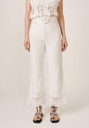 Mujer con pantalones cortos blancos con ribete de encaje, una blusa blanca de encaje floral y sandalias negras con detalles circulares plateados.