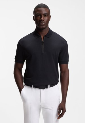 Mand iført en sort kortærmet polo med lynlås, stoppet ned i hvide bukser med sort bælte, stående mod en ensfarvet baggrund.