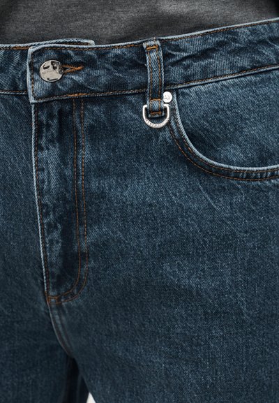 Primer plano de unos vaqueros de mezclilla azul oscuro que muestran un bolsillo delantero, un pasador para el cinturón, un botón metálico de marca y costuras contrastantes en naranja.