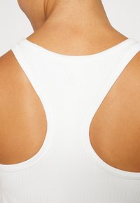 Camiseta sin mangas blanca de canalé con un diseño de espalda nadadora, que presenta un pequeño logo en la parte superior de la espalda. Textura suave, ajuste ceñido y diseño minimalista.