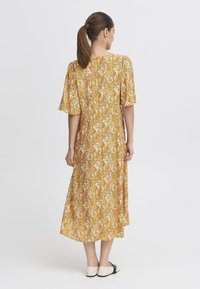 Rochie midi galbenă cu imprimeu paisley, cu mâneci scurte în volan, croi lejer și tiv drept. Modelul poartă pantofi plata de culoare deschisă.