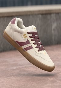 Beige sneaker with maroon stripes and laces, elevated above a textured surface. Gold branding visible on the side and tongue.
