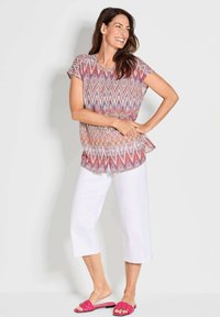 Kurzes, gemustertes Oberteil in Pink, Lila und Weiß, kombiniert mit weißen, cropped Hosen und rosa, gewebten Sandalen.