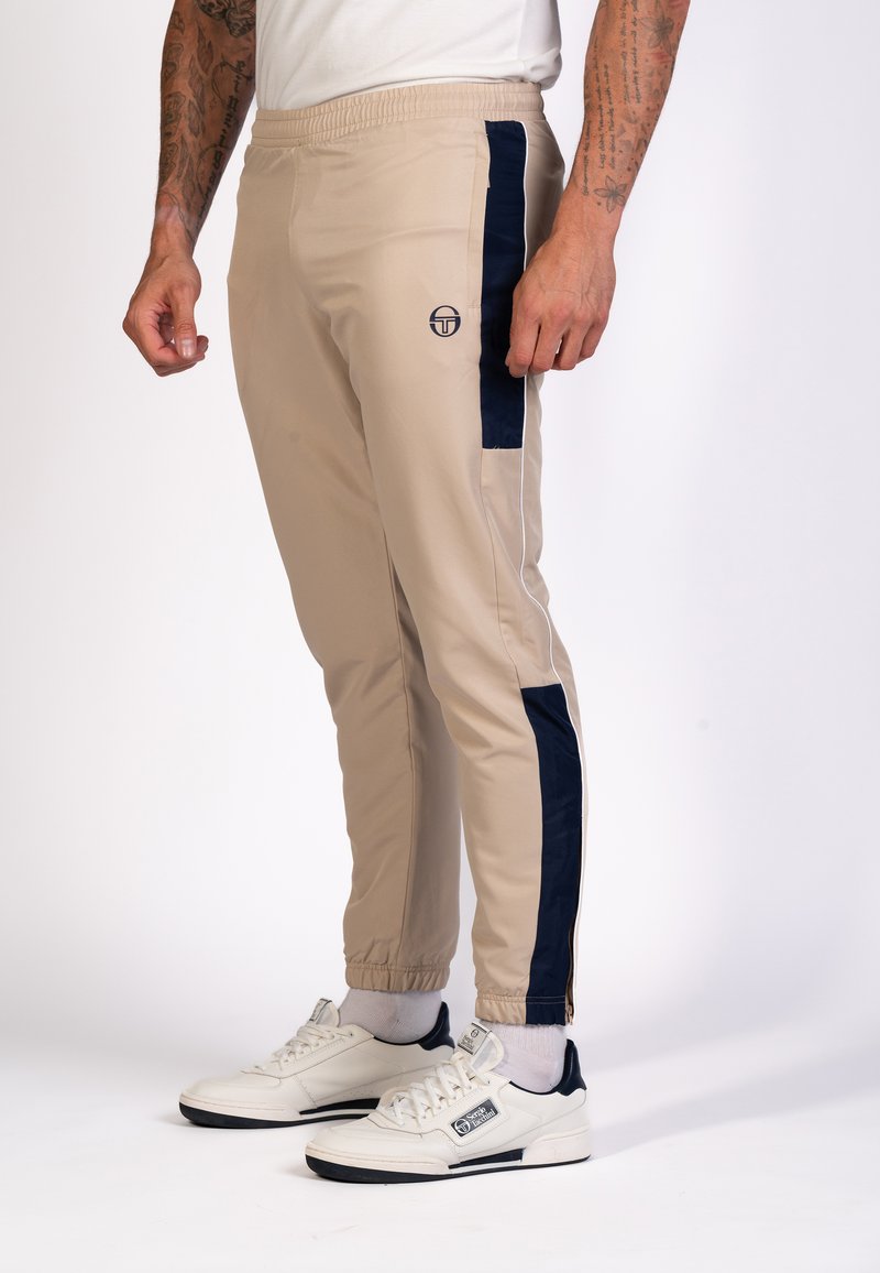 Beige, lichte joggers met marineblauwe zijpanelen, een elastische tailleband en rits sluitingen bij de enkels. Gecombineerd met witte sneakers met marineblauwe accenten.