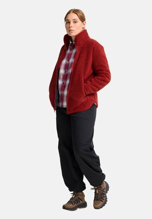 Frau steht und trägt eine rote Fleecejacke, ein kariertes Hemd, schwarze Hose und braune Wanderschuhe vor einem einfarbigen hellen Hintergrund.