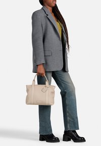 Beige leren tote bag met twee handvatten aan de bovenkant, gestructureerde afwerking en een bagagelabel, gecombineerd met een grijze blazer en denim jeans.