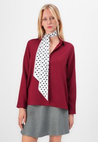 Karmosinröd skjorta med långa ärmar och krage med knappar, i kombination med en grå kjol, accentuerad av en vit scarf med svarta prickar.