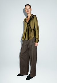 Camicia in satin verde scuro con colletto, abbinata a pantaloni larghi in lana marrone. Modello con scarpe slip-on nere. Silhouette semplice ed elegante.