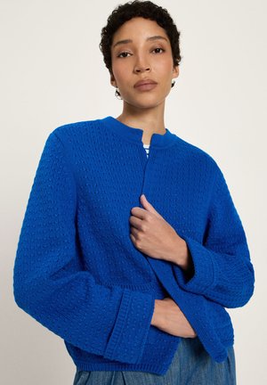 Woman with short curly hair wearing a textured bright blue knit cardigan over jeans, holding the cardigan closed with one hand.