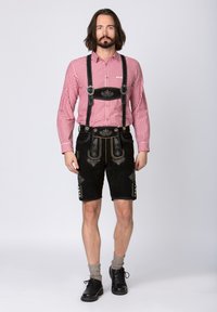 Musta mokkanahka lederhosen, jossa on koristeellinen hopeinen brodeeraus, yhdistettynä punavalkoiseen ruutupaitaan ja harmaisiin polviin ulottuviin sukkiin. Mustat kengät.