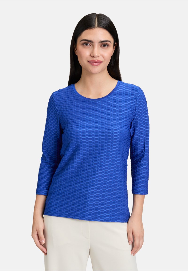 Royalblaue, strukturierte Langarmbluse mit rundem Ausschnitt und gemustertem Stoff, kombiniert mit hellen Hosen.