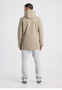 Beige waterproof jacket with a hood, straight cut, and smooth texture, paired with gray sweatpants and white sneakers.