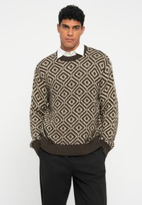 Vīrietis ar melnu un bēšu rombveida adītu džemperi virs baltas kreklu ar apkakli, melnās biksēs, rokas kabatās.