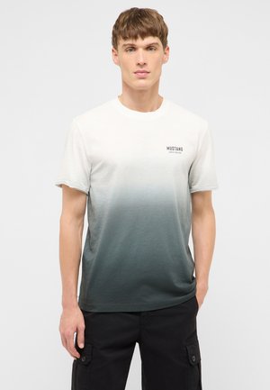 Junger Mann trägt ein kurzärmliges T-Shirt mit einem Farbverlauf von Weiß zu Dunkelgrau und schwarze Cargohosen, steht vor einem schlichten Hintergrund.