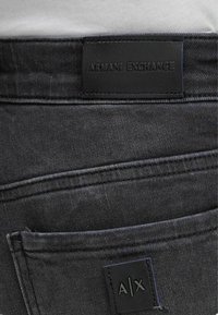 Jeans in denim nero con una finitura testurizzata, dotati di un'etichetta in pelle sulla vita che riporta la dicitura "ARMANI EXCHANGE" e un piccolo cartellino "A/X".