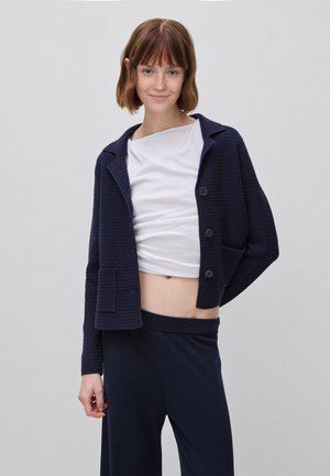 Jonge vrouw met kort bruin haar, gekleed in een marineblauw gebreid vest over een wit cropped topje en wijde marineblauwe broek, staand tegen een grijze achtergrond.