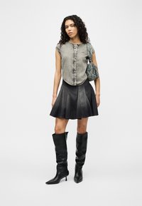 Γυναίκα που φοράει γκρι αμάνικο τοπ από τζιν, μαύρη φούστα με φουσκωτή γραμμή, μαύρες μπότες ως το γόνατο και κρατάει μια μπλε ραφιασμένη τσάντα ώμου.