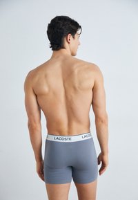 Grijze boxershorts met een witte tailleband met het "LACOSTE" logo. Glad textuur en aansluitend ontwerp, weergegeven vanuit een achteraanzicht.