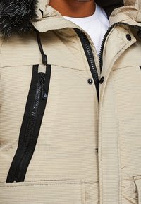 Homme portant une veste d'hiver beige avec des détails zippés noirs, une capuche en fourrure, et une chemise blanche en dessous, visible du cou jusqu'au milieu du torse.