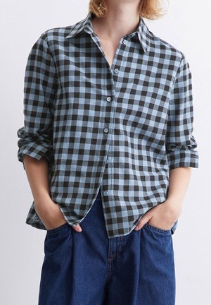 Persoon draagt een blauw-zwart geruit overhemd met opgerolde mouwen en donkerblauwe spijkerbroek met handen in de zakken.