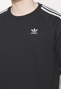 Svart Adidas-tröja med vita tre-stripes detaljer på axlarna och liten vit Adidas trefoil-logga på bröstet.