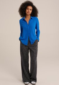 Camicia blu con bottoni e maniche lunghe, abbinata a pantaloni grigi a gamba larga con motivo, e calzature di colore chiaro. Design semplice e vestibilità comoda.