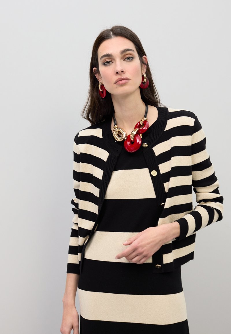 Mustavalkoraidallinen neuletakki, jossa pyöreä pääntie ja kullansävyiset napit, kerrostettuna yhteen raidallisen mekon kanssa. Rohkea punainen kaulakoru ja korvakorut.