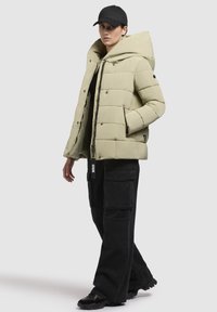 Giacca a vento in verde oliva chiaro, design trapuntato con ampio cappuccio, chiusura con zip e bottoni a pressione. Abbinata a pantaloni cargo neri e berretto.