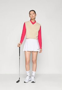 Mulher em pé com um taco de golfe, vestindo um colete sem mangas bege sobre uma camisa vermelha, saia branca, meias brancas e tênis brancos contra um fundo simples.