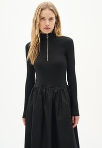 Rochie neagră cu nervuri, cu gât înalt și detaliu cu fermoar, având un corset ajustat și o fustă plisată fluidă, confecționată dintr-un material neted.