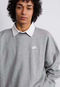 Siv sivka z ohlapnim ovratnikom, ki ima majhen bel Nike logotip na prsih. Tkanina se zdi mehka in teksturirana, sproščen kroj.