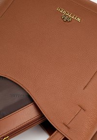 Bruine leren handtas met een gestructureerde afwerking. Voorzien van een gouden logo aan de voorkant en een ritsluiting. Binnenin is er een voering met logo-patroon.