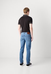 Jauns vīrietis stāv ar muguru, ģērbies melnā polo kreklā, gaiši zilās džinsās ar brūnu ādas uzlīmi un melnbaltās sporta apavos.