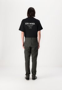 Osoba o brązowych włosach do ramion, stojąca tyłem, ubrana w czarną koszulkę ESS.WRD., oliwkowe spodnie i czarne trampki z białymi paskami.