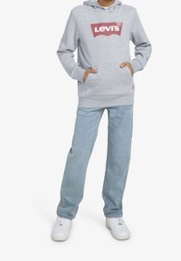 Felpa grigia con cappuccio con il logo "LEVI'S", tasche frontali, abbinata a jeans azzurri e sneaker bianche. Vestibilità rilassata e design casual.