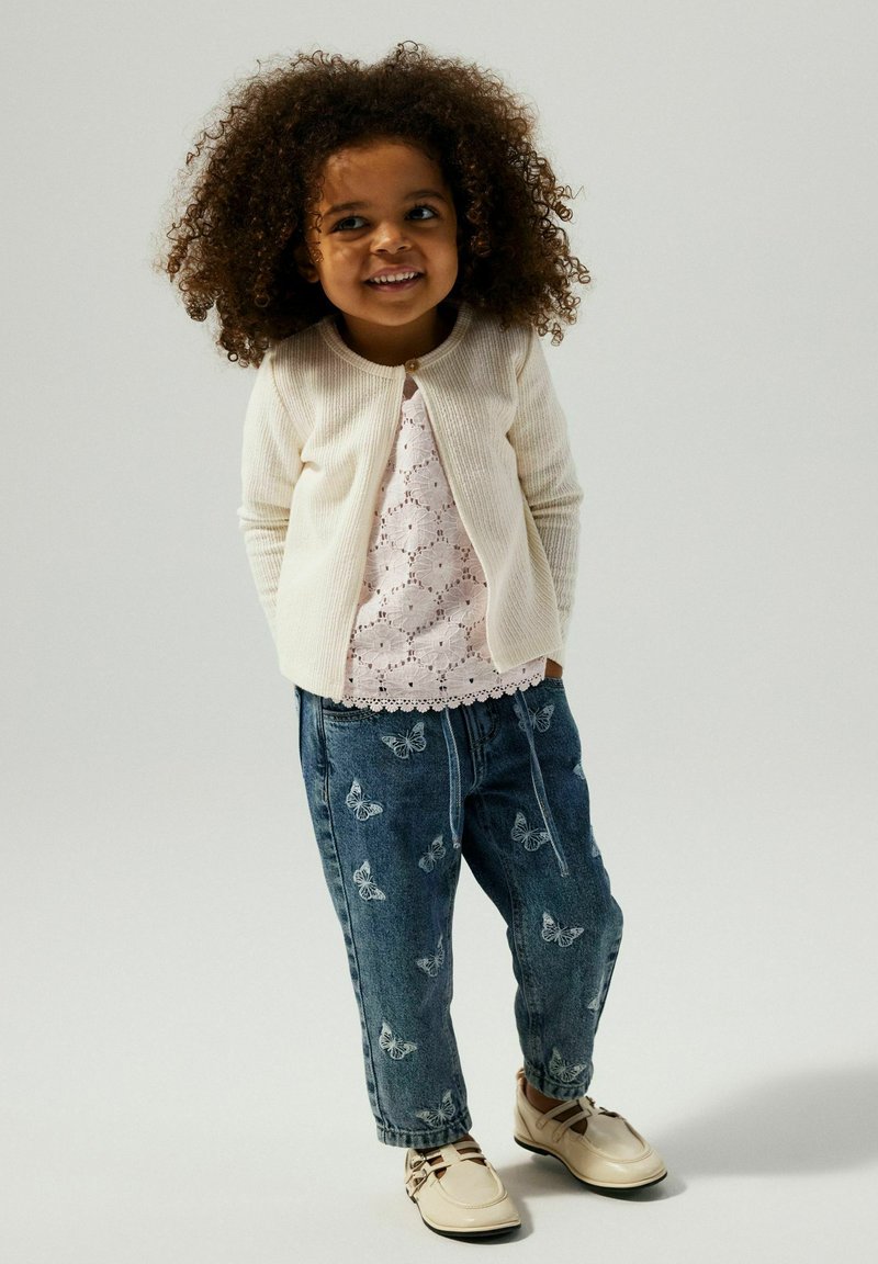 Cardigan crema, top in pizzo rosa, jeans blu con motivo a farfalla e scarpe beige. Presenta materiali testurizzati ed elementi di design giocosi.