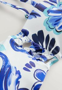 Kostium kąpielowy w kwiaty w kolorze białym, z niebieskimi i turkusowymi abstrakcyjnymi wzorami. Materiał gładki z marszczeniami oraz regulowanymi ramiączkami.