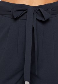 Pantalons taille haute de couleur bleu marine foncé avec une ceinture en tissu nouée en nœud, présentant des plis et une texture lisse.