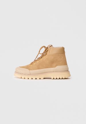 Botte montante en daim beige avec semelle en caoutchouc côtelée et embout, col rembourré et fermeture à lacets sur fond uni.