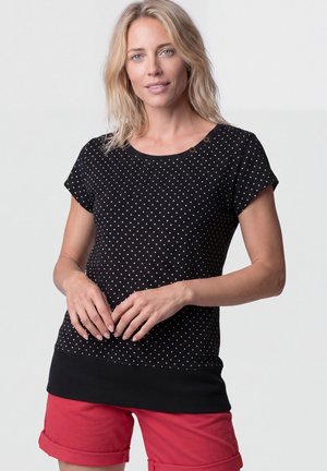 Blonde woman wearing black short-sleeve polka dot top and red cuffed shorts, standing with hands gently clasped at waist.