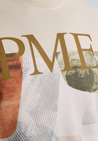 Krémové bavlněné tričko s velkým zlatým nápisem "PME". Design obsahuje geometrické vzory v hnědé, šedé a zelené barvě. Měkká textura.