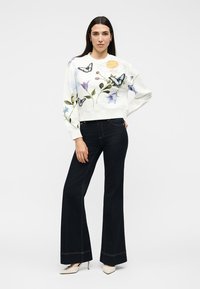 Femme portant un sweat-shirt blanc avec des papillons et des fleurs colorés, associé à un pantalon large noir taille haute et des talons blancs.