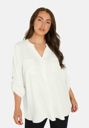 Vrouw met lang bruin haar, die een losse witte blouse met opgerolde mouwen en een zwarte broek draagt, staand tegen een effen achtergrond.