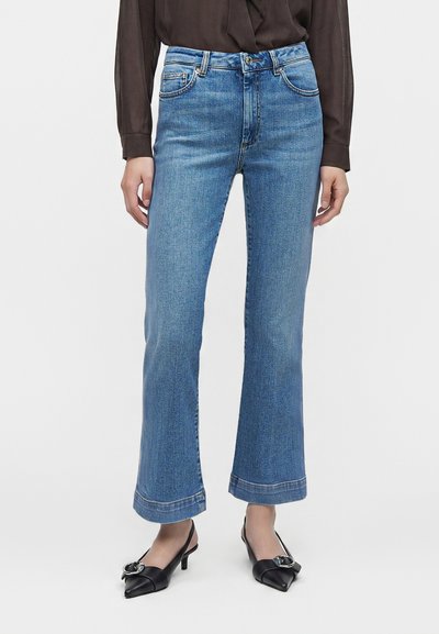 Femme portant un jean droit taille haute, lavage moyen, un chemisier noir transparent à manches longues et des talons pointus noirs avec boucles.