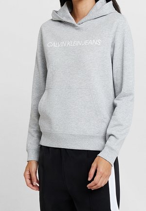 Henkilö, joka käyttää vaaleanharmaata Calvin Klein Jeans -hupparia, jossa on etutasku, ja mustia housuja, joissa on valkoinen sivuraita.