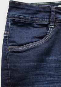 Jeans de mezclilla azul oscuro con un bolsillo frontal visible, costuras en amarillo contrastante y una textura suave. Cuenta con un pasador en la cintura.